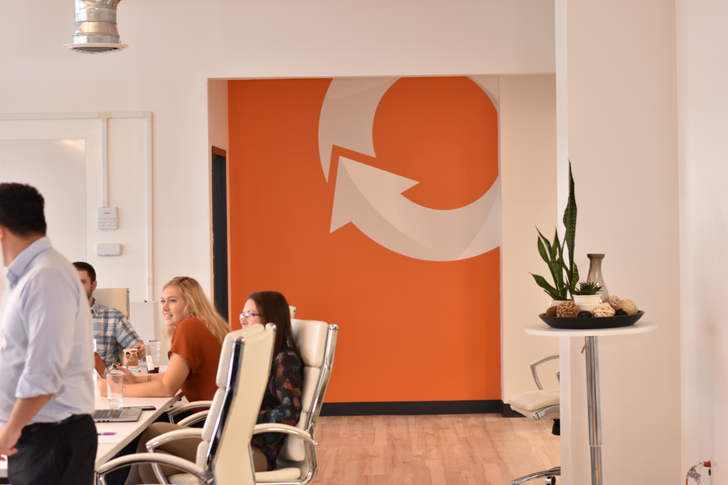 Employees sitting for a meeting in an office with orange wall and white logo.