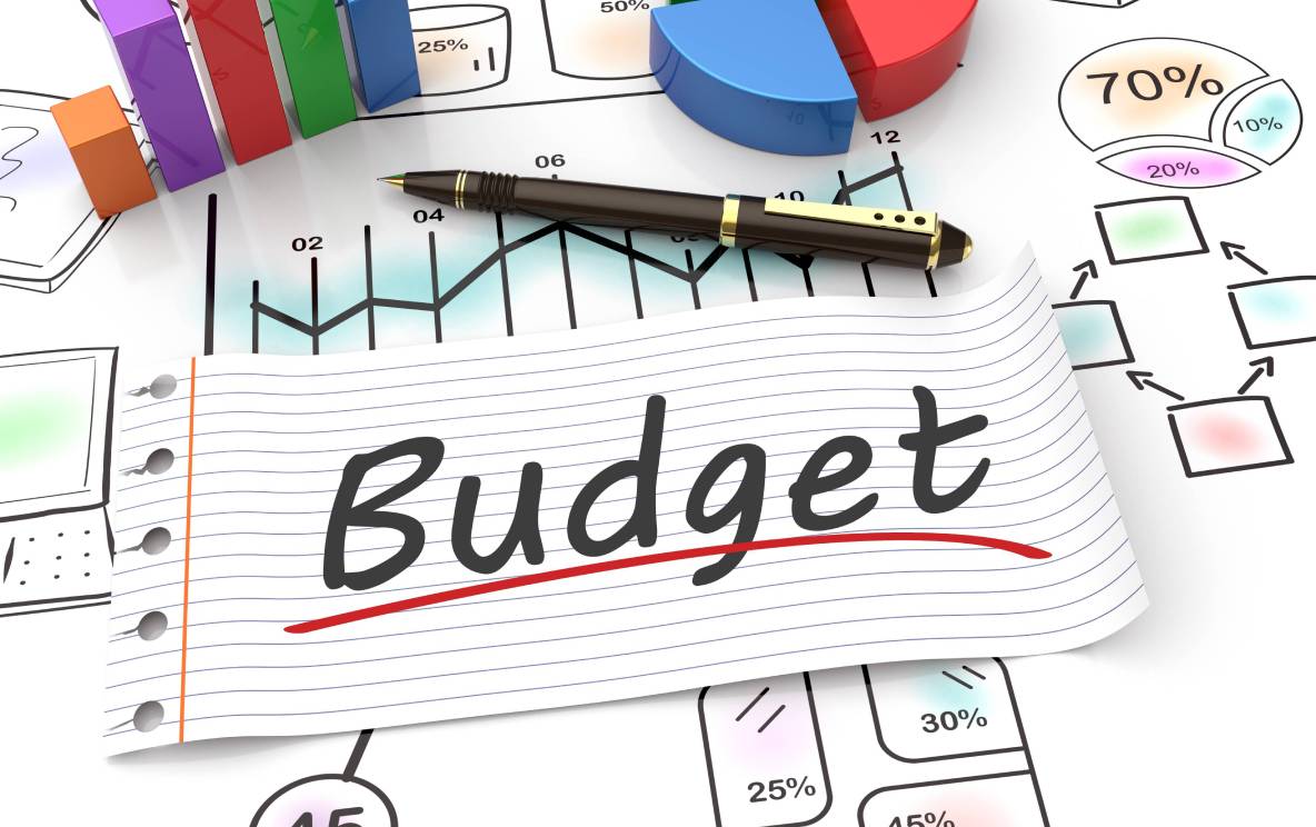 The word “budget” is written on a piece of paper, lying on a desk amidst calculations, graphs, and expense projections.