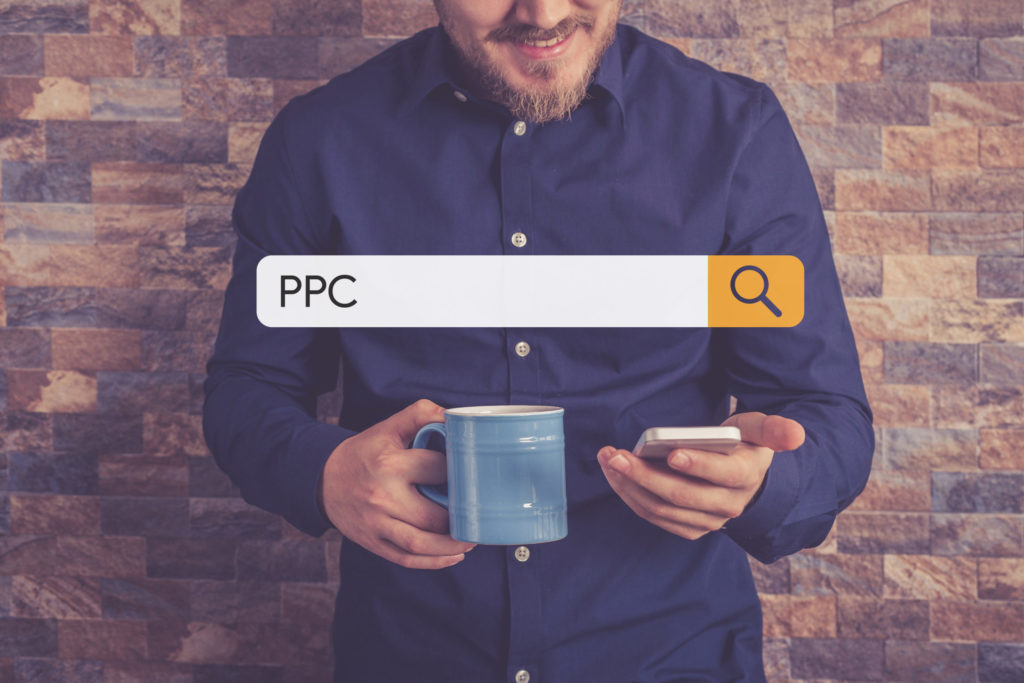 Man in blue shirt holding a coffee mug smiles as he looks down at his phone with “PPC” in the search bar. 