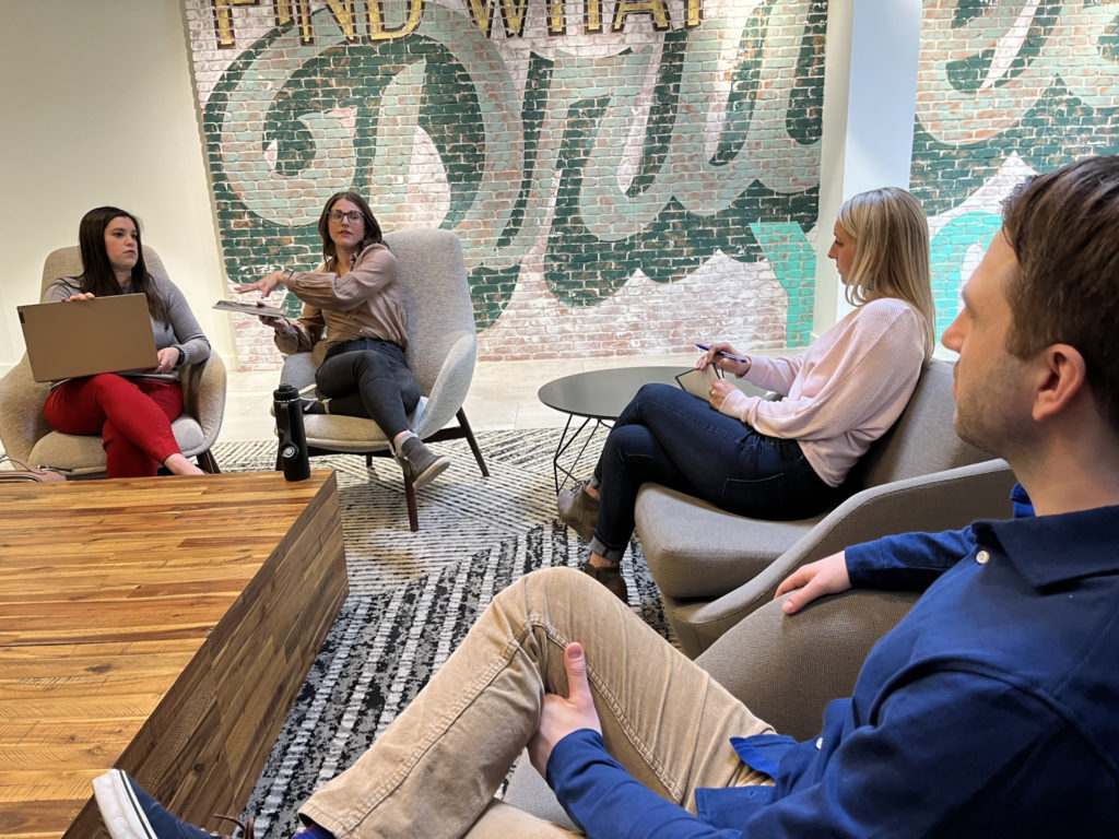 Team of marketing professionals having a discussion in gray chairs with brick accent wall in background. 