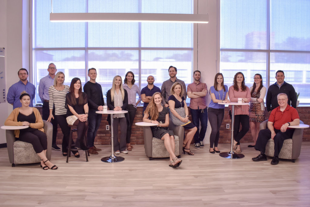 The employees of a marketing agency pose for a group photo in front of large windows.