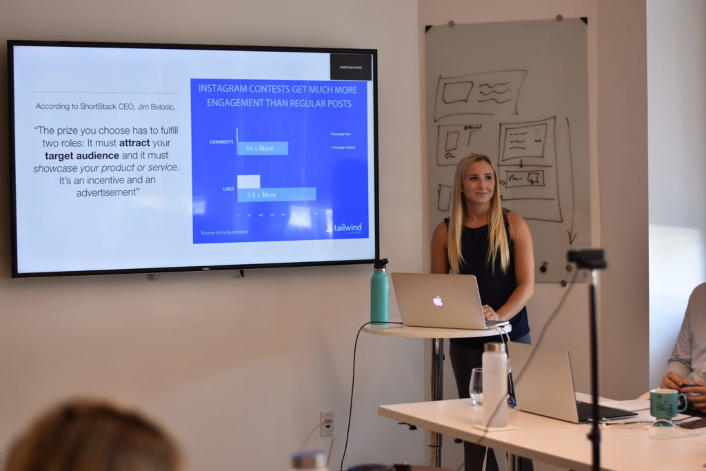 A blonde woman stands at a podium with her laptop while delivering a presentation to a group of people.