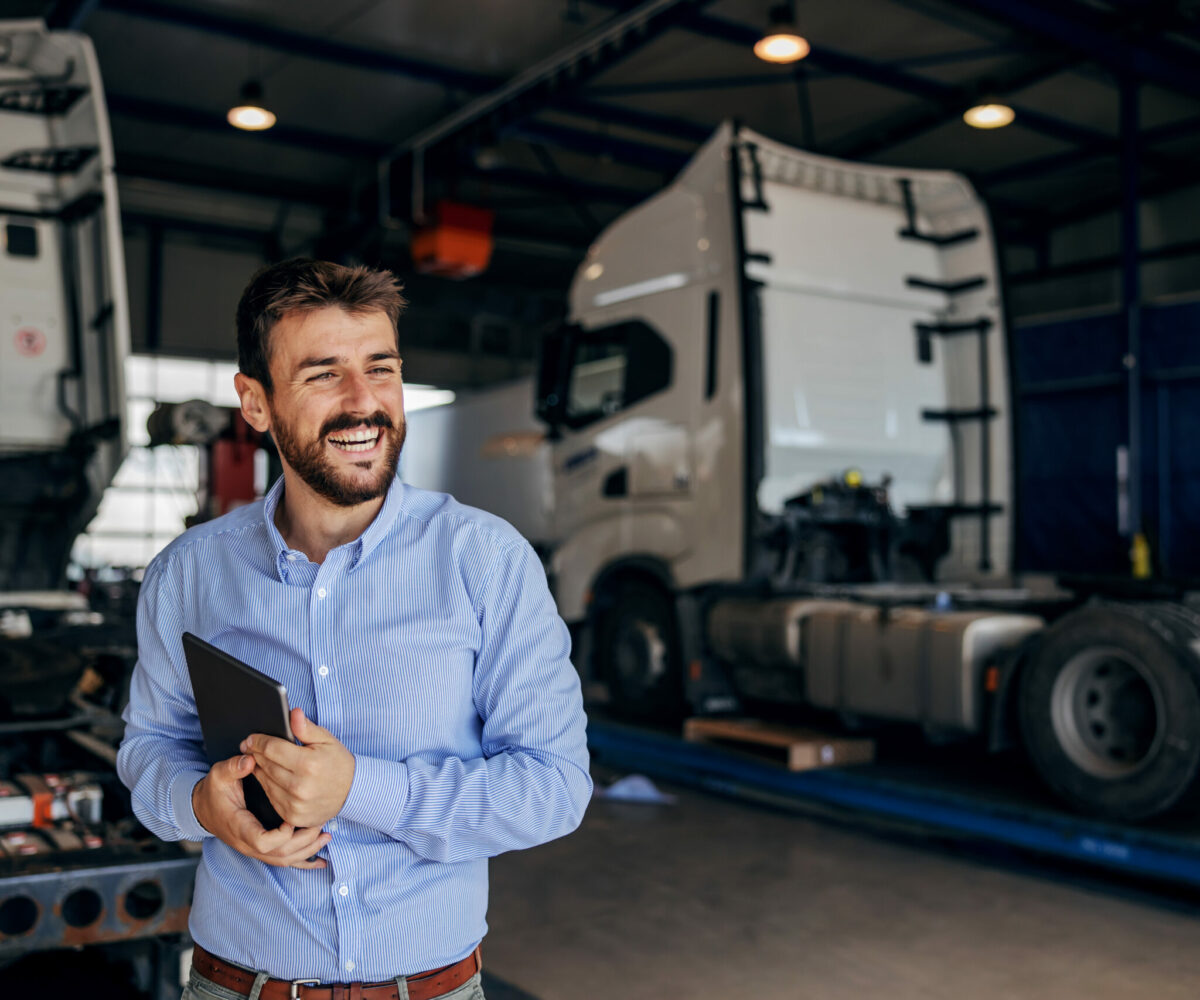  Smiling manufacturing executive in blue shirt holding tablet with trucks in the background.